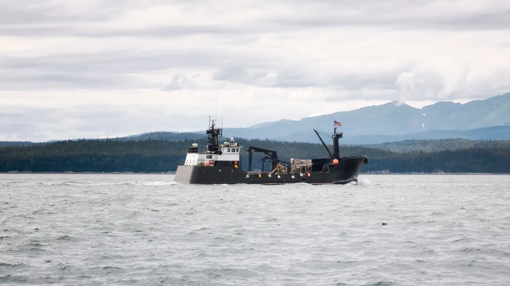 A trawler underway on grey water off a forested Alaska coast